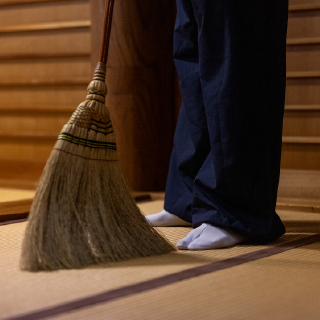 Close-up photo of a broom near the feet of someone sweeping tatami flooring while wearing traditional Japanese clothing.