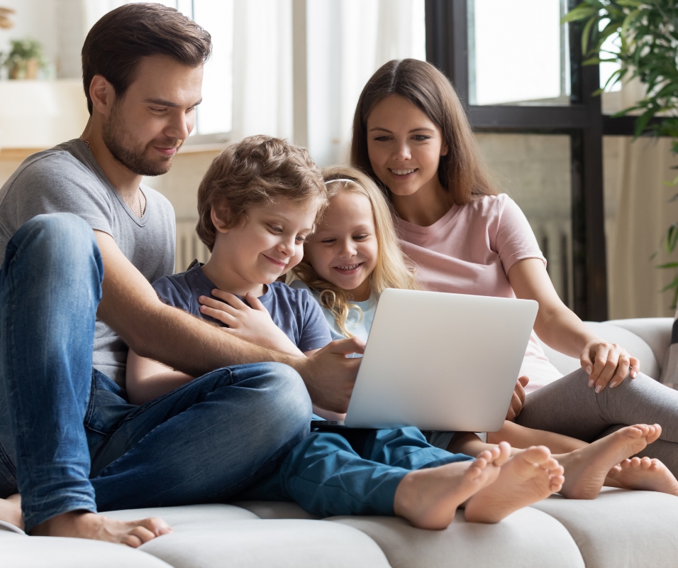 Gambar anak-anak melihat PC sambil bersantai di ruang keluarga bersama orang tua duduk di dekat mereka.