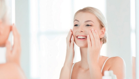 An image of moisturing, a woman smiling touching her face in front of the mirror.