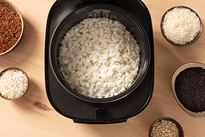 Vista superior de la olla arrocera SR-DA152KXE en color negro, llena de arroz cocido y rodeada de pequeños tazones que contienen diferentes tipos de arroz.
