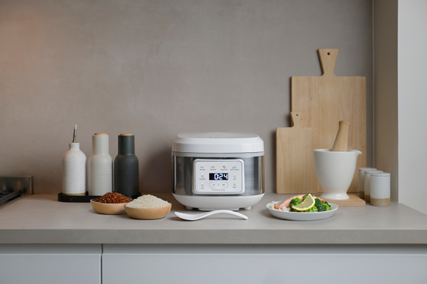 Olla arrocera SR-DA152WXE en color blanco colocada sobre una encimera de cocina, rodeada de tablas de cortar, utensilios de cocina y varios ingredientes.