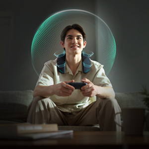 A person sits on a couch wearing a neck speaker while playing video games.