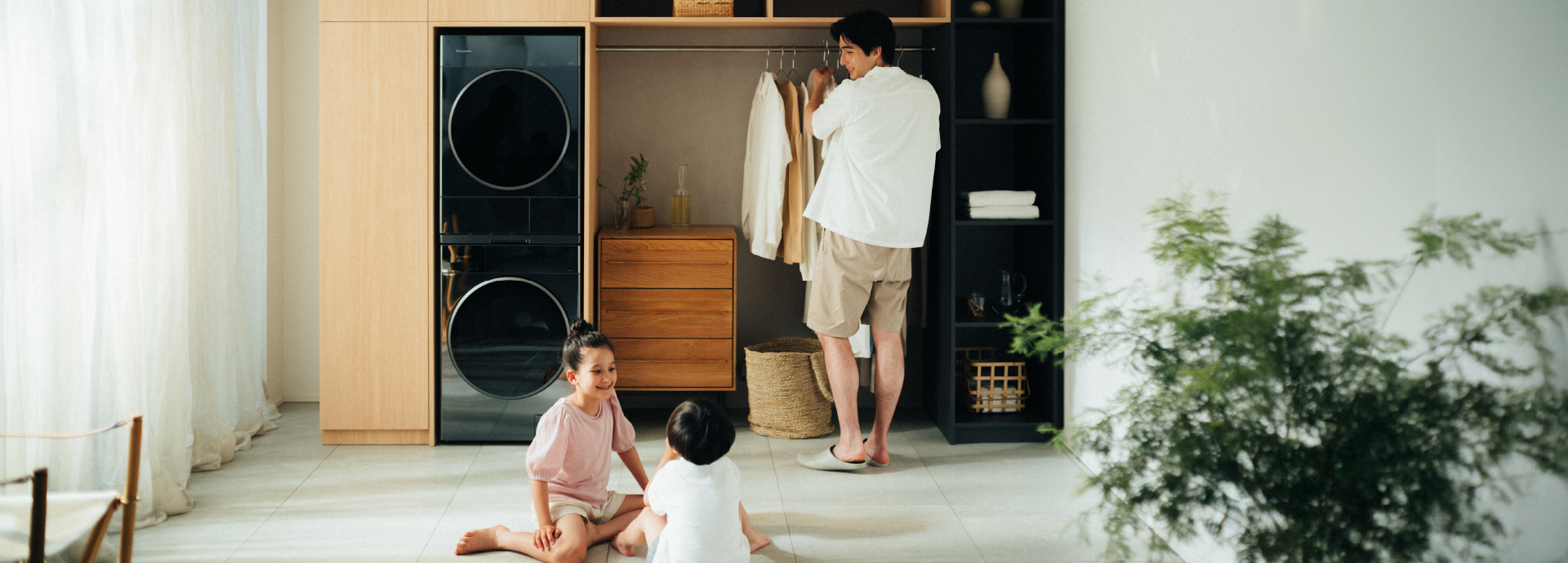 Section Banner: Photo of two stacked washer-dryers beautifully integrated with a minimalist furniture unit with closets and shelving in a bright, open room. A man is hanging clothes while two young children play on the floor nearby.
