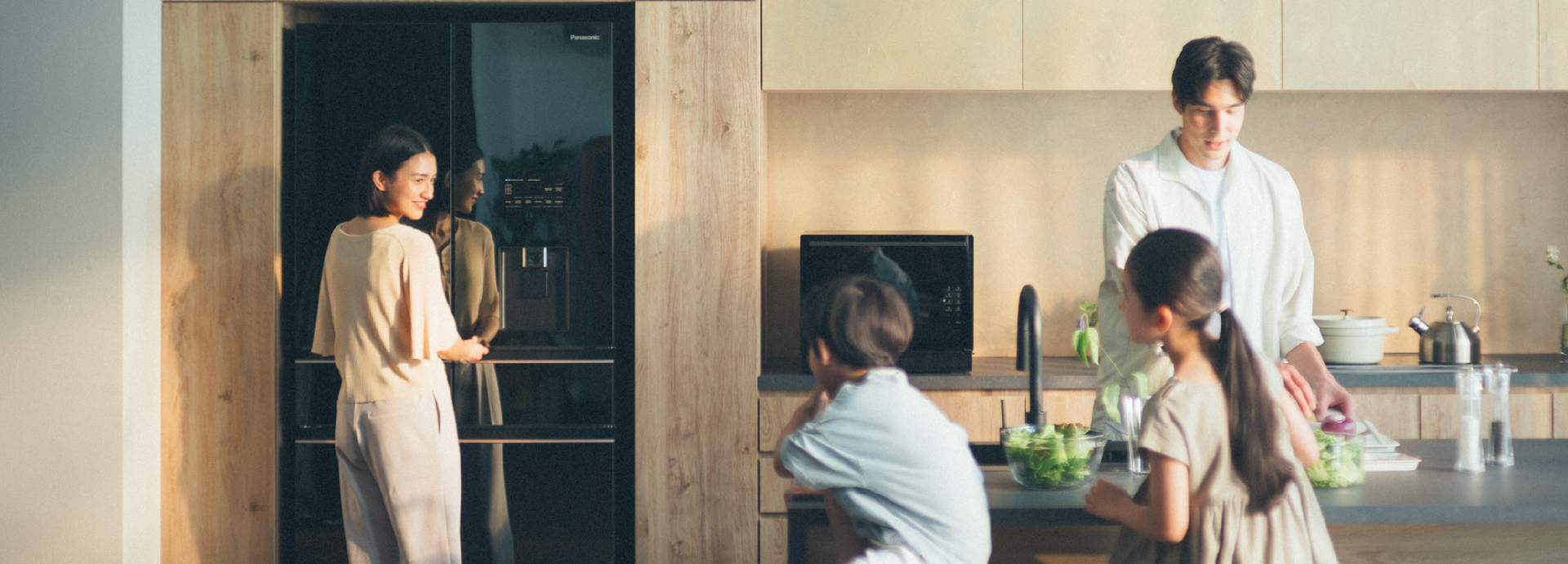 Family in a bright, modern kitchen. The mother smiles at her kids as she opens a Panasonic refrigerator while the father preps fresh ingredients.