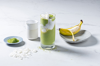 Un pahar înalt cu smoothie de matcha și cocos, cu un model în straturi de verde și alb, decorat cu fulgi de cocos și un praf de pudră de matcha. În jurul paharului se află o banană decojită pe o farfurie gri, un recipient mic alb cu lapte de cocos, un vas albastru cu pudră de matcha și fulgi de cocos presărați pe o suprafață de marmură albă.
