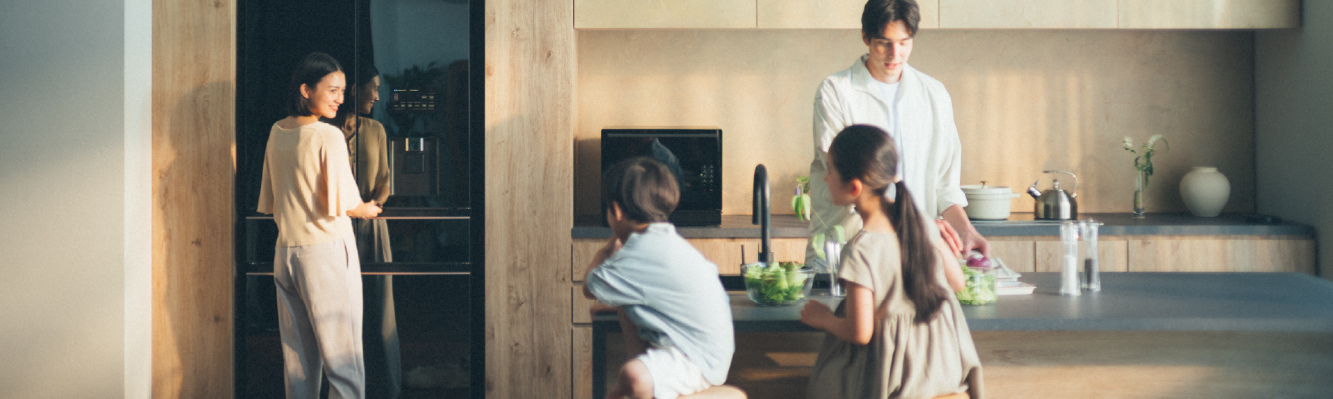 Top Banner: Photo of a family in a bright and warm minimalist kitchen. The mother is standing by a stylish, mirror-finish refrigerator. She is smiling at two young children sitting at a kitchen island across from their father who is preparing food.
