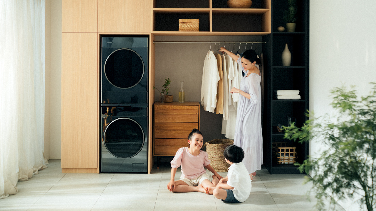 A mother hanging clothes in a closet near 2 stacked washer-dryers while smiling at kids playing in a bright room.