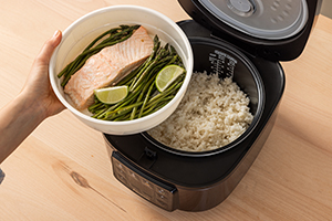 Top view of the SR-DA152KXE Rice Cooker in Black filled with rice, with a hand holding a plate of salmon, vegetables, and spices, and the Water Indicator visible.