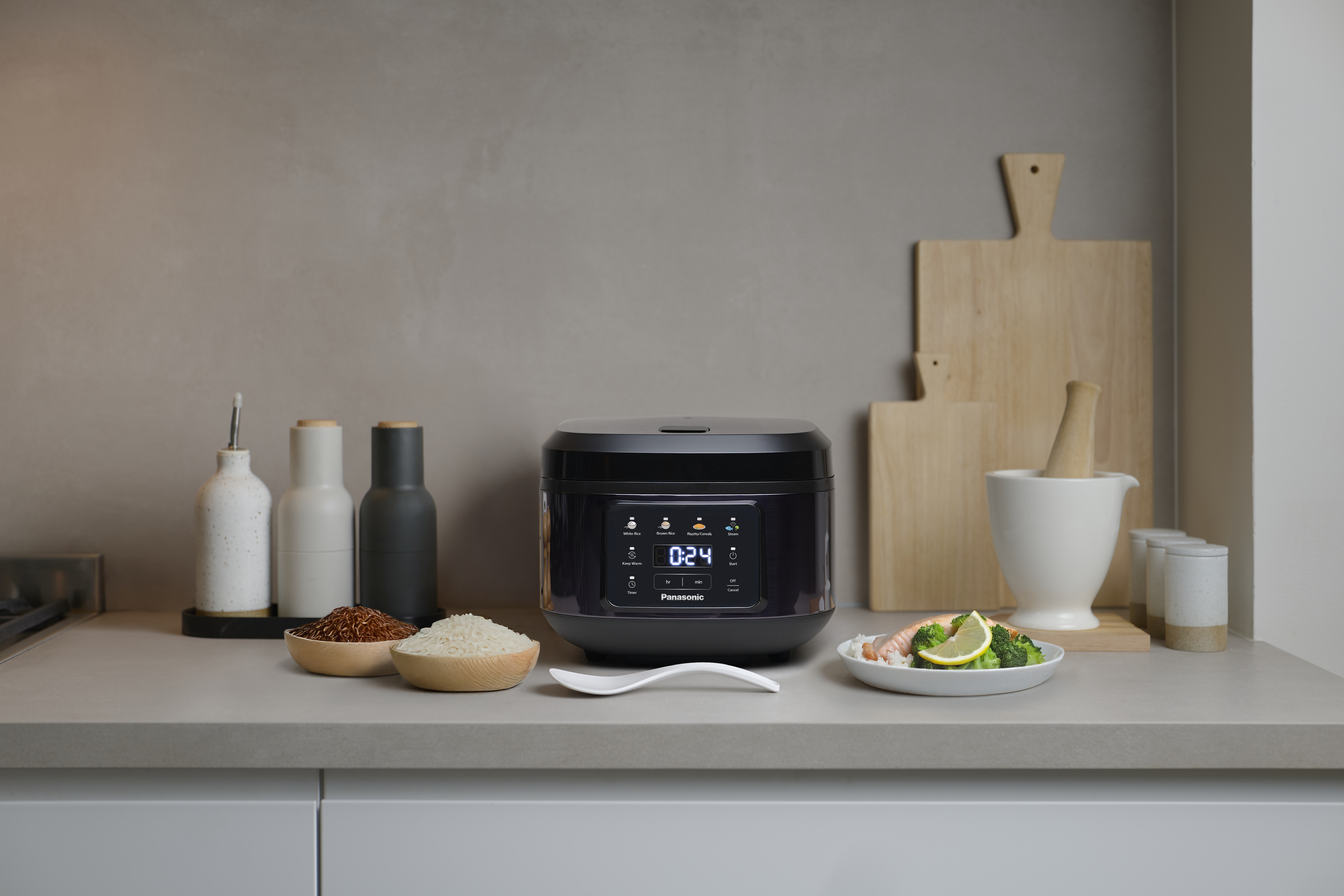 SR-DA152KXE Rice Cooker in Black placed on a kitchen counter, surrounded by cutting boards, cooking utensils, and various ingredients.