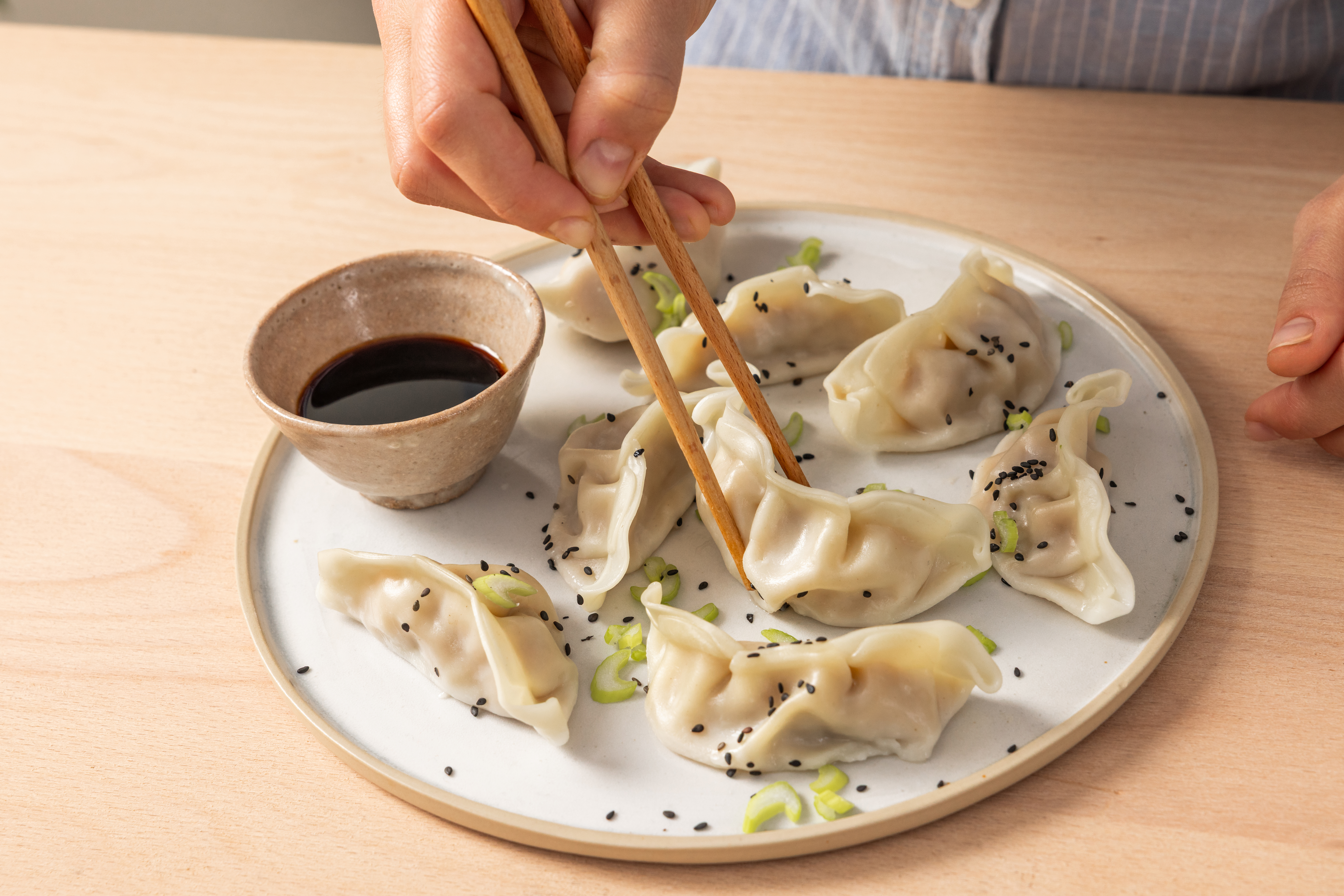 A hand using chopsticks to pick up dumplings from a plate with dipping sauce.