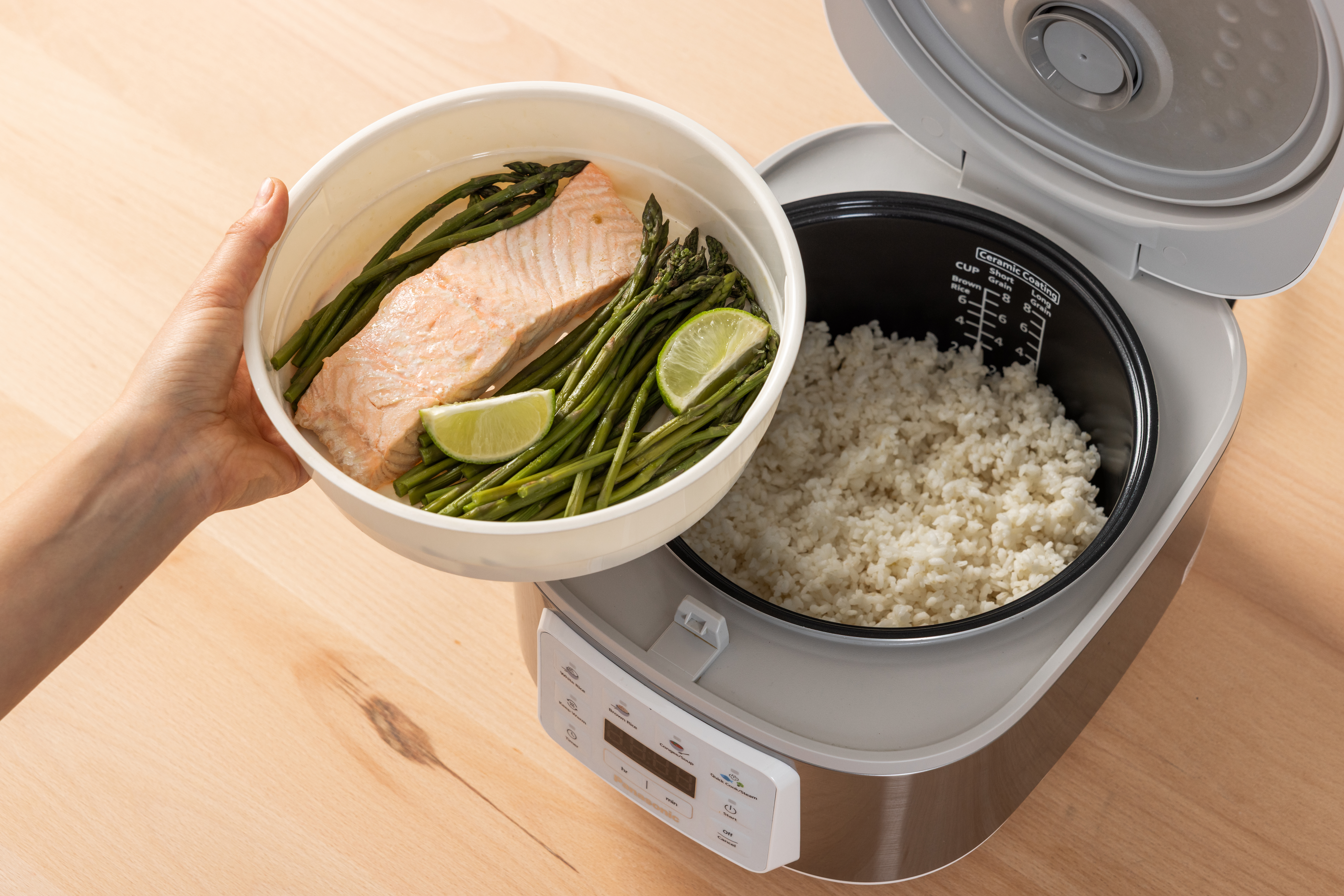 Top view of the SR-DA152WXE Rice Cooker in White filled with rice, with a hand holding a plate of salmon, vegetables, and spices, and the Water Indicator visible.