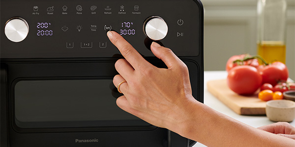 Et nærbillede af en hånd, der justerer indstillingerne på Panasonic Flex Air Fryer NF-BC1000. Et skærebræt med tomater, grøntsager og en olieflaske er synlige i baggrunden, hvilket viser et køkkenmiljø.