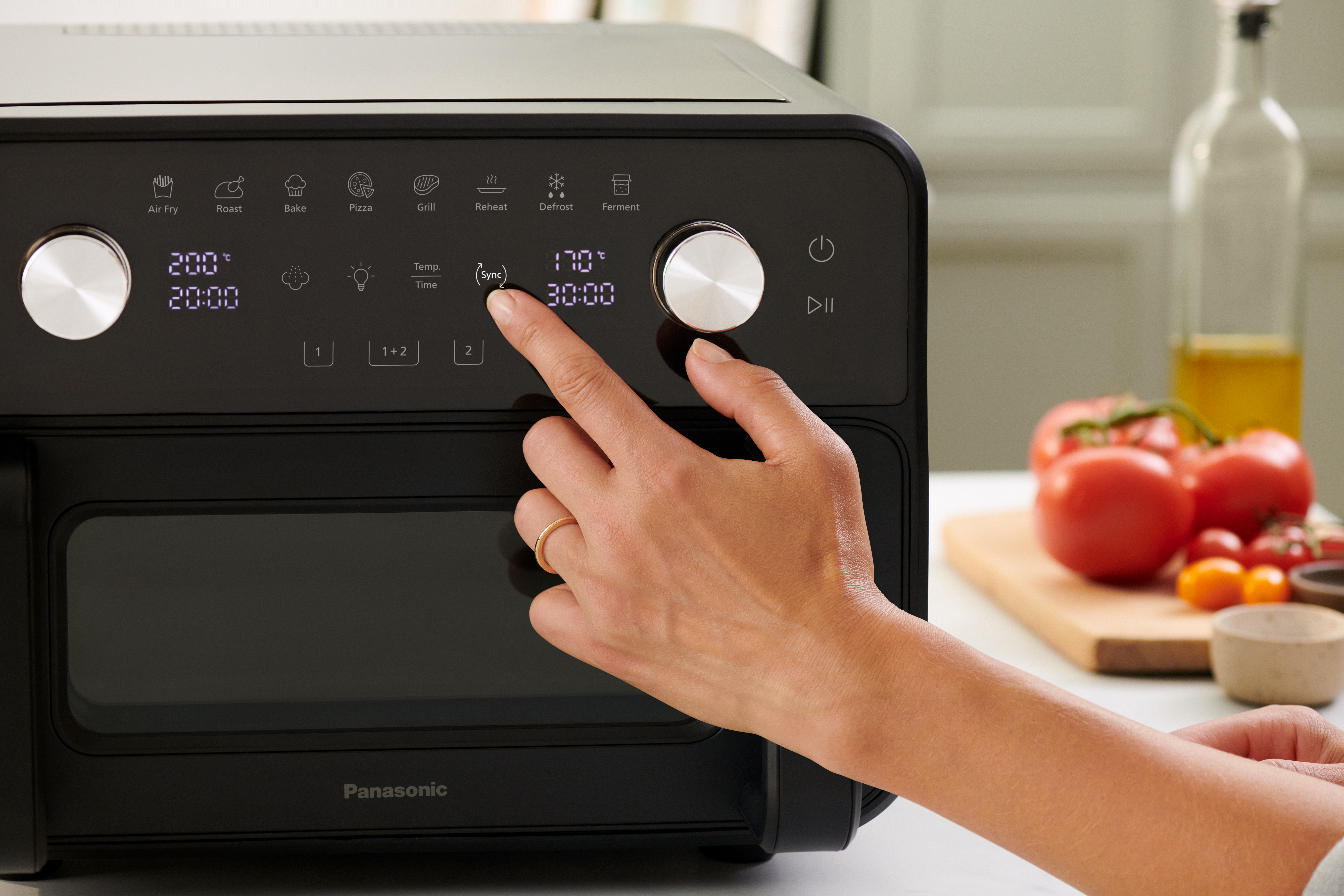 Et nærbillede af en hånd, der justerer indstillingerne på Panasonic Flex Air Fryer NF-BC1000. Et skærebræt med tomater, grøntsager og en olieflaske er synlige i baggrunden, hvilket viser et køkkenmiljø.