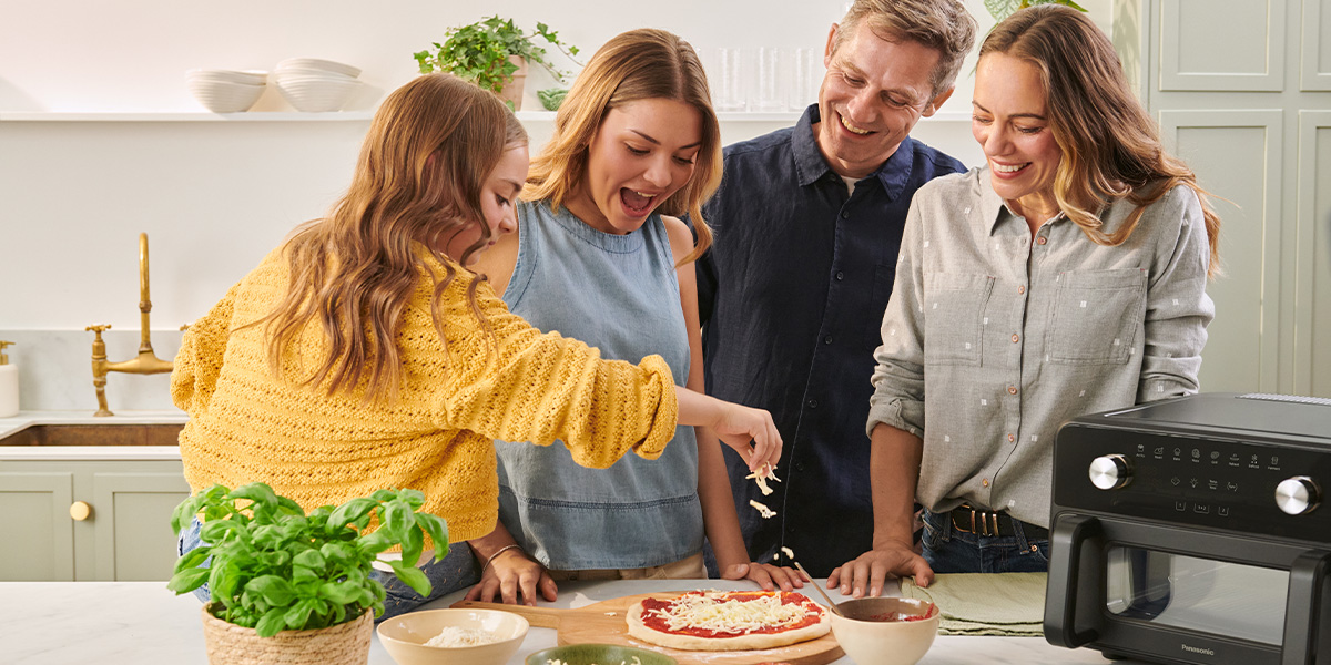 En familie samles omkring køkkenbordet og laver en pizza sammen. En person smører ost på pizzaen, mens de andre ser på og smiler, mens de foreviger et fælles madlavningsøjeblik i et hyggeligt hjemmekøkken.