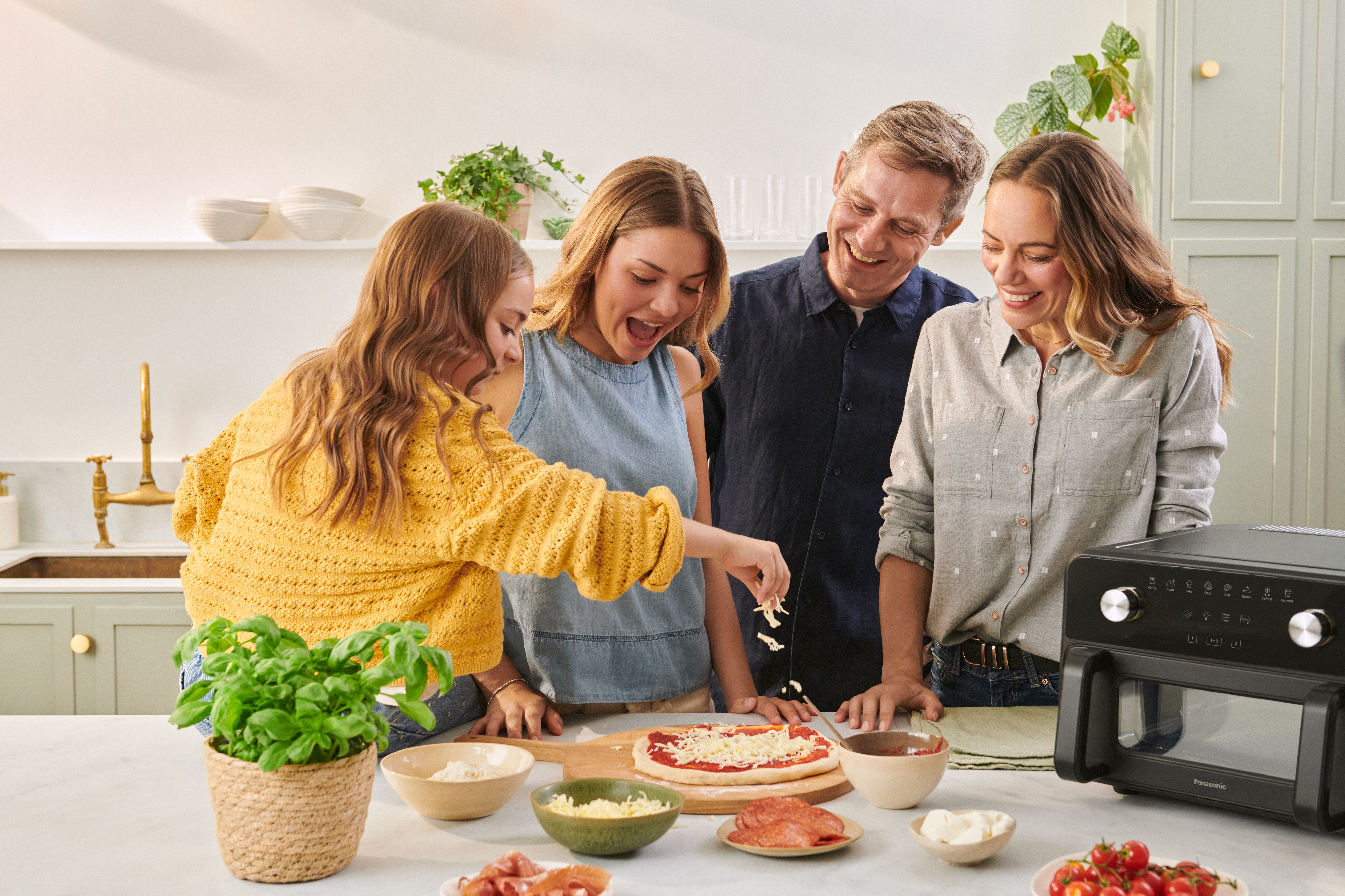En familie samles omkring køkkenbordet og laver en pizza sammen. En person smører ost på pizzaen, mens de andre ser på og smiler, mens de foreviger et fælles madlavningsøjeblik i et hyggeligt hjemmekøkken.