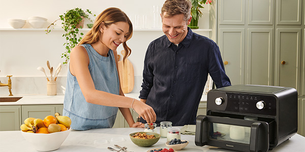 To personer tilbereder mad sammen i et hjemmekøkken. Den ene tilsætter ingredienser til en skål, mens den anden ser på og smiler. Bordpladen har skåle, krukker, frugt og Panasonic Flex Air Fryer NF-BC1000, hvilket skaber en hyggelig madlavningsscene.