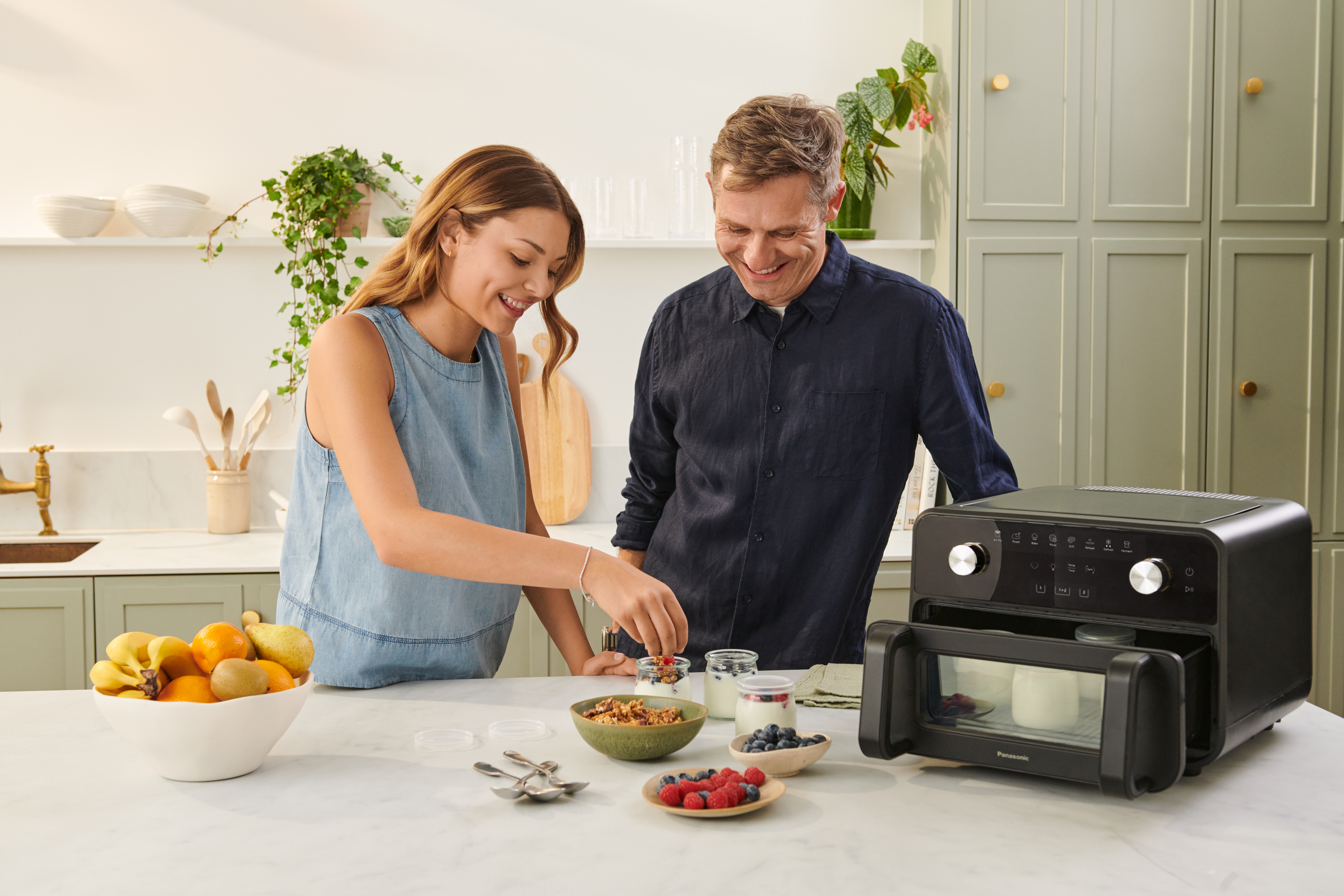 To personer tilbereder mad sammen i et hjemmekøkken. Den ene tilsætter ingredienser til en skål, mens den anden ser på og smiler. Bordpladen har skåle, krukker, frugt og Panasonic Flex Air Fryer NF-BC1000, hvilket skaber en hyggelig madlavningsscene.