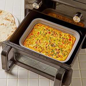 En gryde med en bagt blanding af æg og grøntsager placeres inde i Panasonic Flex Air Fryer NF-BC1000 med døren åben. En tortilla eller et fladbrød ligger på køkkenbordet ved siden af apparatet.