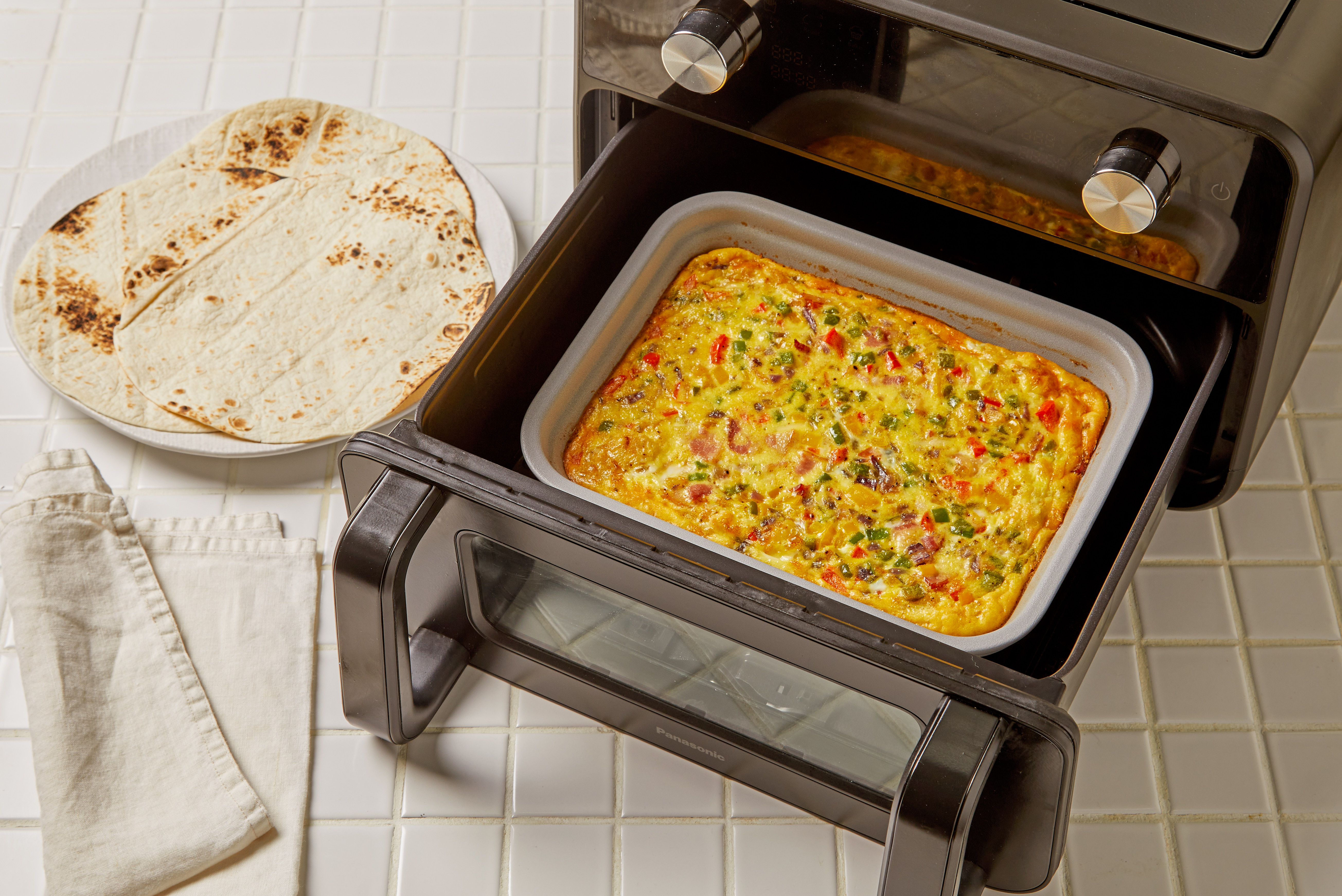 En gryde med en bagt blanding af æg og grøntsager placeres inde i Panasonic Flex Air Fryer NF-BC1000 med døren åben. En tortilla eller et fladbrød ligger på køkkenbordet ved siden af apparatet.