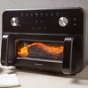 Panasonic Flex Air Fryer NF-BC1000 placeret på køkkenbordet med mad, der tilberedes indeni, og oplyst af det indvendige lys.