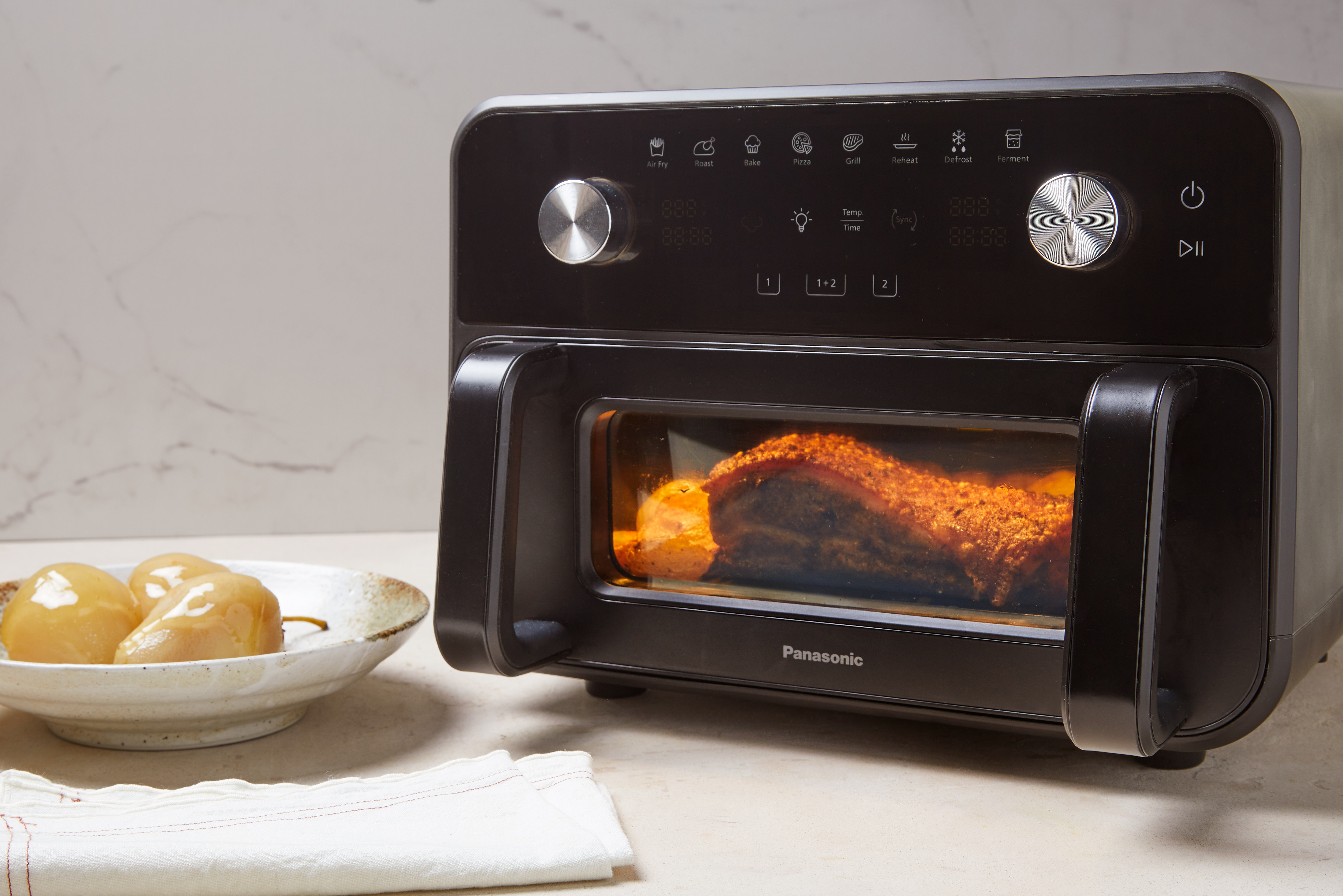 Panasonic Flex Air Fryer NF-BC1000 placeret på køkkenbordet med mad, der tilberedes indeni, og oplyst af det indvendige lys.