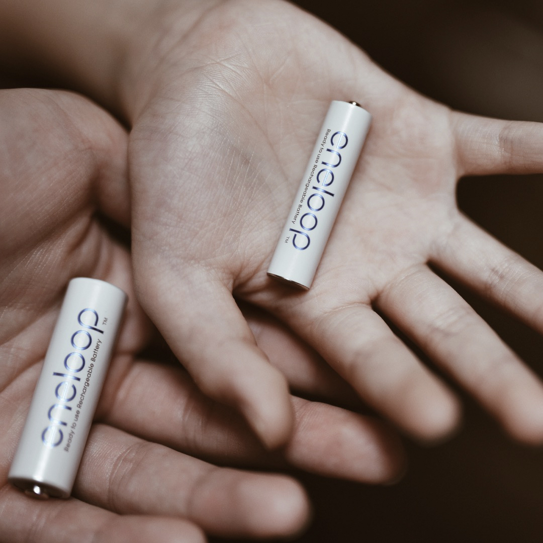 An adult's hand holds an eneloop AA battery in its palm; a child's hand holds an eneloop AAA battery.