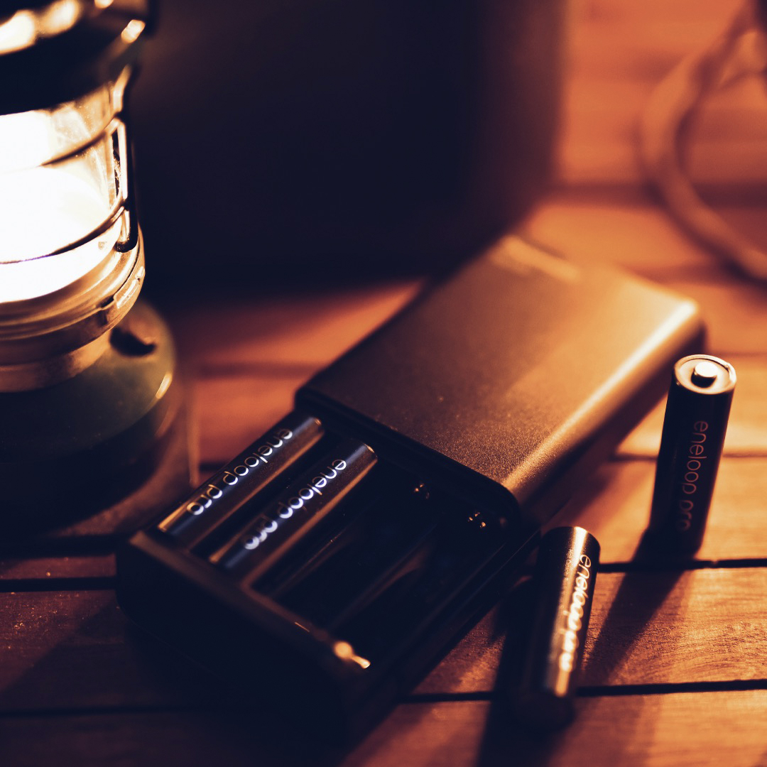 A charger rests on a wooden floorboard next to a battery-powered camping lantern. Two batteries are in the charger, two lay beside it.