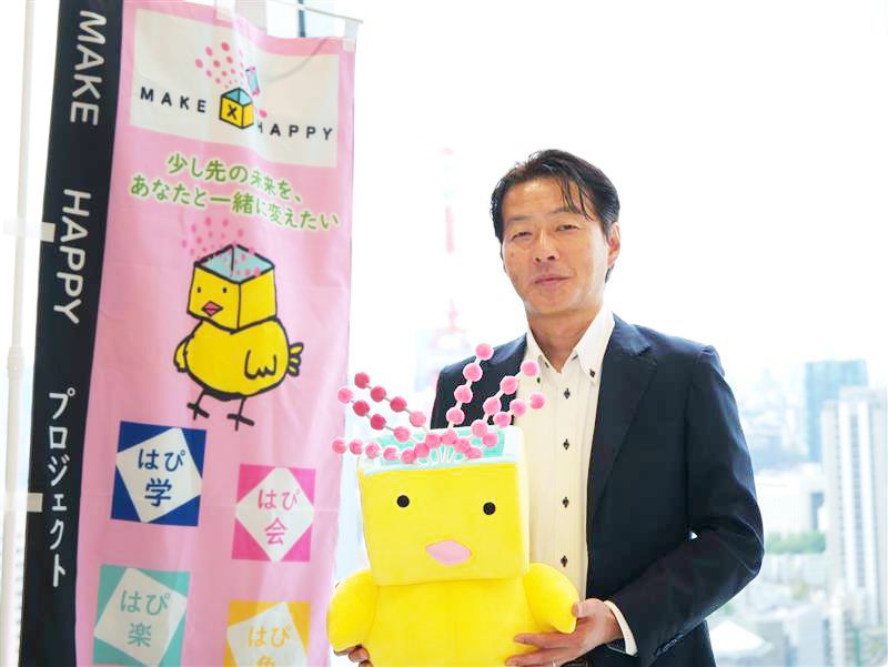 パナソニック インダストリーの小澤社長が「はっぴーさん®」のぬいぐるみを持ち、プロジェクト名入りののぼり旗の横に立つ様子。