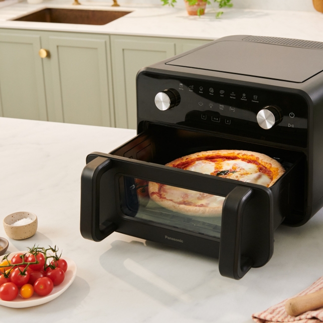 The Panasonic Flex Air Fryer NF-BC1000 is shown on kitchen countertop with its big basket open, with a pizza inside. Cherry tomatoes and salt are placed nearby.