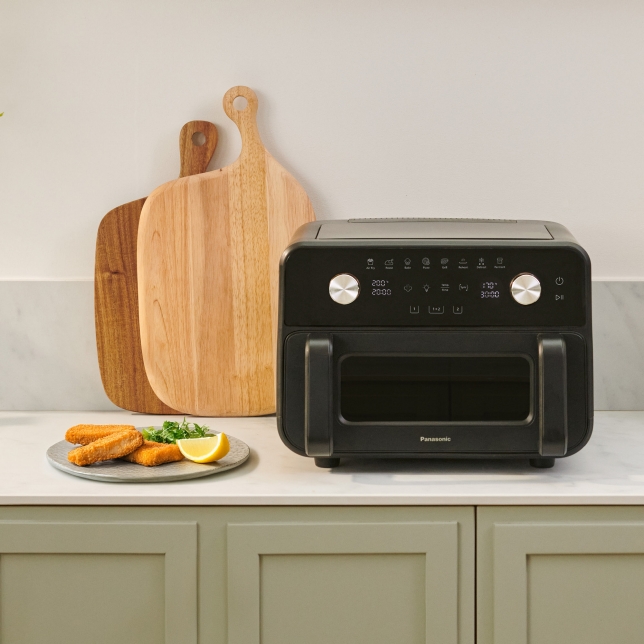 The Panasonic Flex Air Fryer NF-BC1000 is placed on a kitchen counter with cutting boards behind it and next to a plate with food.