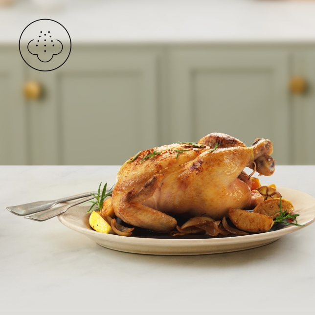 A roasted chicken served on a white plate with lemon slices and herbs, set in a kitchen counter. An icon in the top left corner highlights the Gentle Steam Technology used by the Panasonic Flex Air Fryer NF-BC1000.