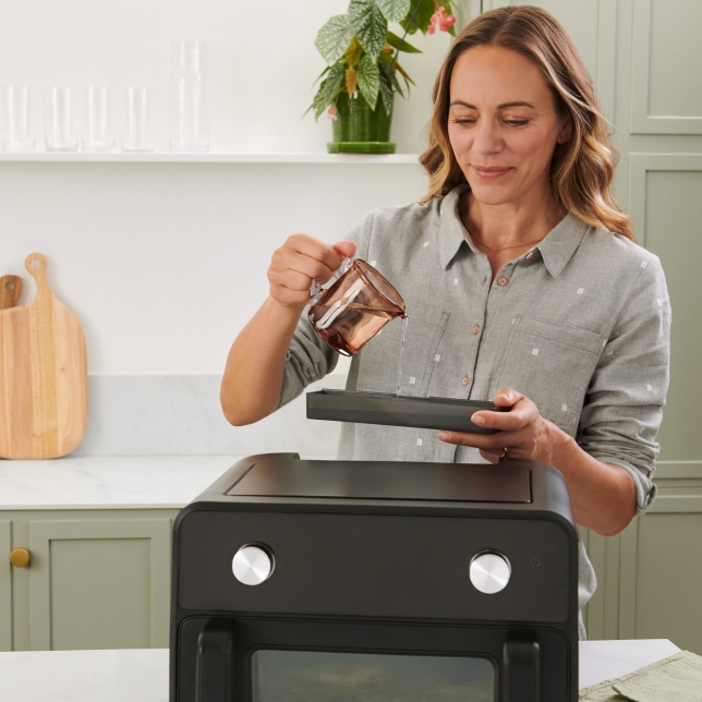 A woman pours water into the integrated tank of the Panasonic Flex Air Fryer NF-BC1000. The appliance is placed in a modern kitchen setting.