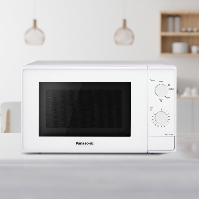 A white Panasonic NN-E20JWMEBG microwave on a kitchen counter. The background shows a modern kitchen with pendant lights and shelves holding various items.