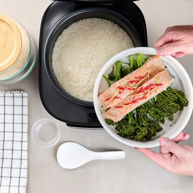 Top view of the SR-DA152KXE Rice Cooker in Black filled with rice, with a hand holding a plate of salmon, vegetables, and spices.