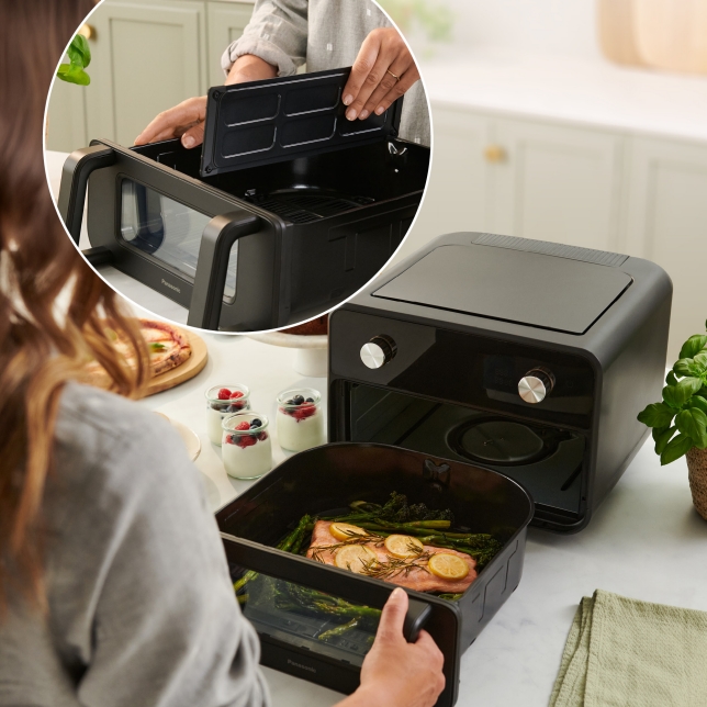 Een persoon opent de mand van de Panasonic Flex Air Fryer NF-BC1000, die op een aanrecht staat. In de grote mand zijn zalm en groenten zichtbaar. In de linkerbovenhoek is een andere afbeelding te zien van een persoon die de scheider vasthoudt die de grote mand in twee kookzones verdeelt.