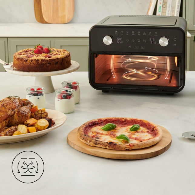 De Panasonic Flex Air Fryer NF-BC1000 staat op een aanrecht in de keuken, met een grafische weergave van de 360° Air Flow Technology. Rondom het apparaat liggen een pizza, geroosterd vlees met groenten, taarten en yoghurtpotjes.