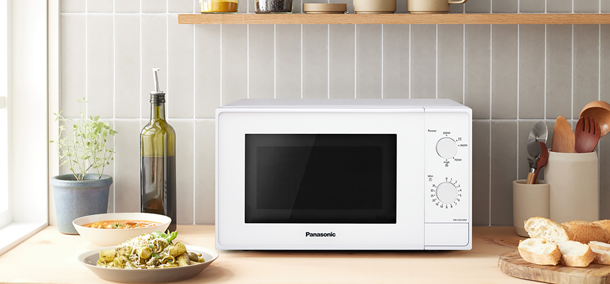 Front view of the Panasonic NN-E20JWMEBG microwave with the door closed, placed on a kitchen counter. Surrounding it are lifestyle elements such as a bottle of olive oil, a bowl of uncooked pasta, and a wooden spoon, suggesting a cozy cooking environment.