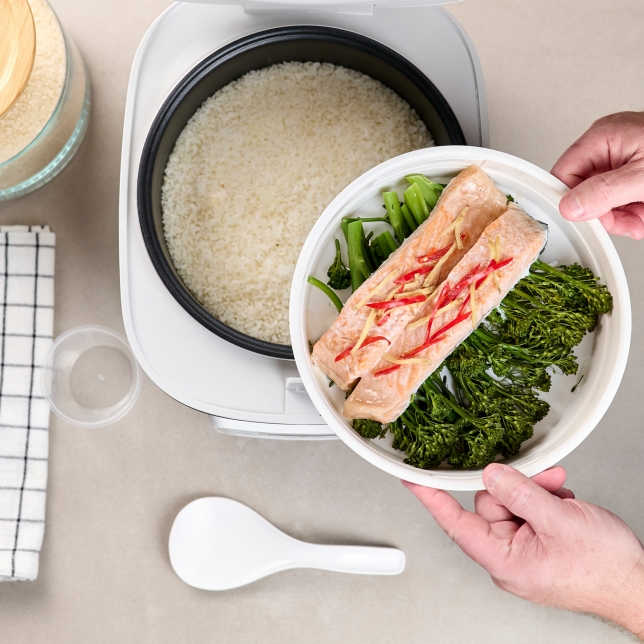 Top view of the SR-DA152WXE Rice Cooker in White filled with rice, with a hand holding a plate of salmon, vegetables, and spices.