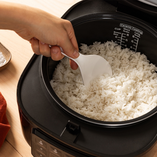 Vista superior de la arrocera SR-DA152KXE en color negro, donde se observa una mano extrayendo arroz cocido con la cuchara medidora desde el interior del aparato. Se aprecia el indicador de agua.