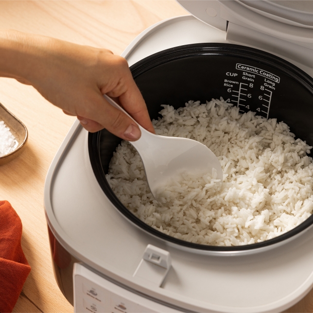 Vista superior de la arrocera SR-DA152WXE en color blanco, donde se observa una mano extrayendo arroz cocido con la cuchara medidora desde el interior del aparato. Se aprecia el indicador de agua.