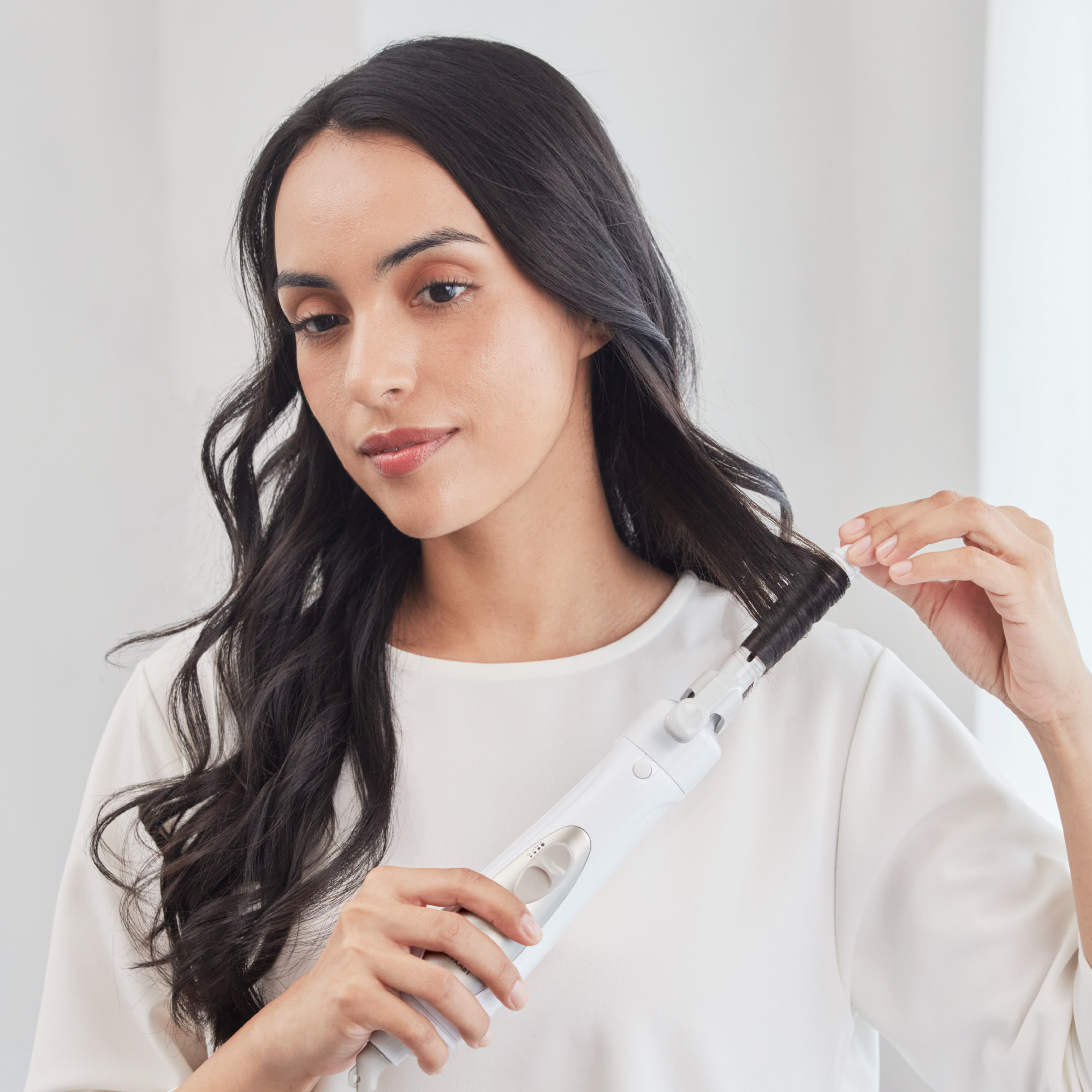 A woman with long, dark, wavy hair using a hair styler to curl a section of her hair.