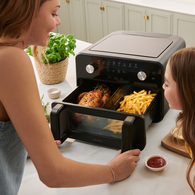 Panasonic Flex Air Fryer NF-BC1000 med korgen öppen. Två flickor kontrollerar kyckling och pommes frites i de två synliga tillagningszonerna.